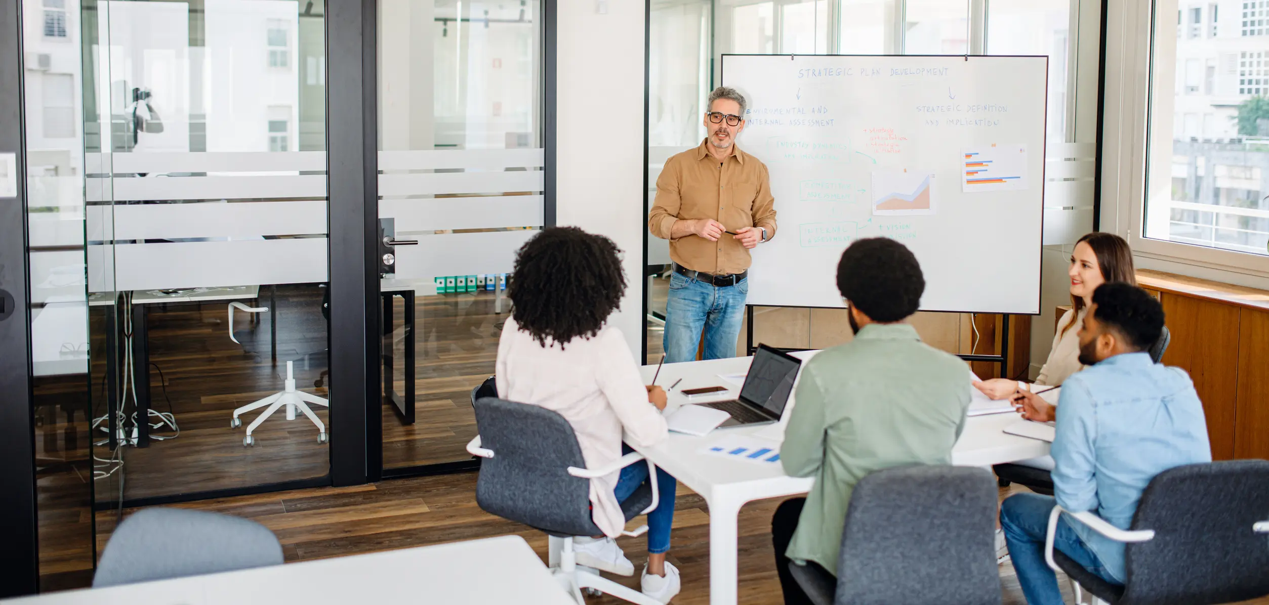 Focused team engages in strategic planning session in modern office. The attentive group surrounds a whiteboard as an experienced leader outlines the workflow, exemplifying a collaborative atmosphere
